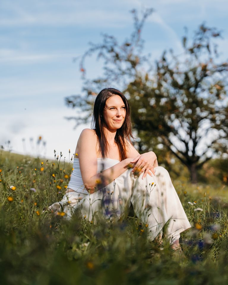 Nelia sitzt lächelnd in einer Blumenwiese, hinter ihr Obstbäume und blauer Himmel.