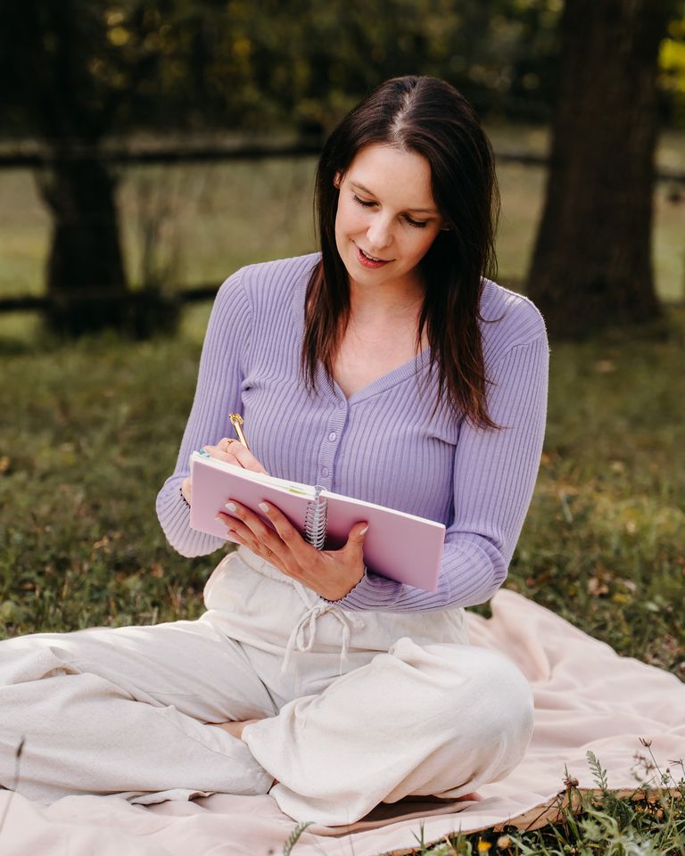 Nelia sitzt auf einer Decke im Gras und schreibt in ein Notizbuch.