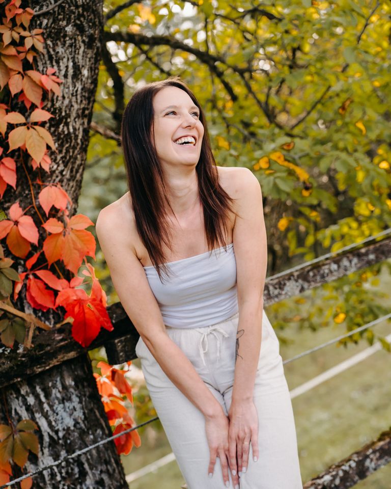 Nelia lacht herzlich, sie lehnt an einem Zaun vor Herbstlaub.