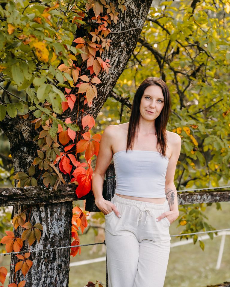 Nelia lehnt lächelnd an einem Holzzaun, neben ihr ein Baumstamm mit rotem Herbstlaub.